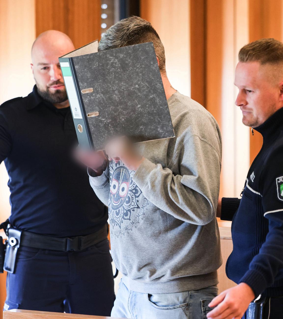Der angeklagte Krankenpfleger hält sich einen Aktenordner vor sein Gesicht, während er von Justizangestellten zu seinem Platz gebracht wir (Archivfoto)