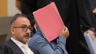 Auf dem Bild zu sehen ist: Einer der drei Angeklagten sitzt zwischen Anwälten in einem Gerichtssaal des Hochsicherheitstrakts des Oberlandesgerichts und verdeckt sein Gesicht hinter einem Aktendeckel.