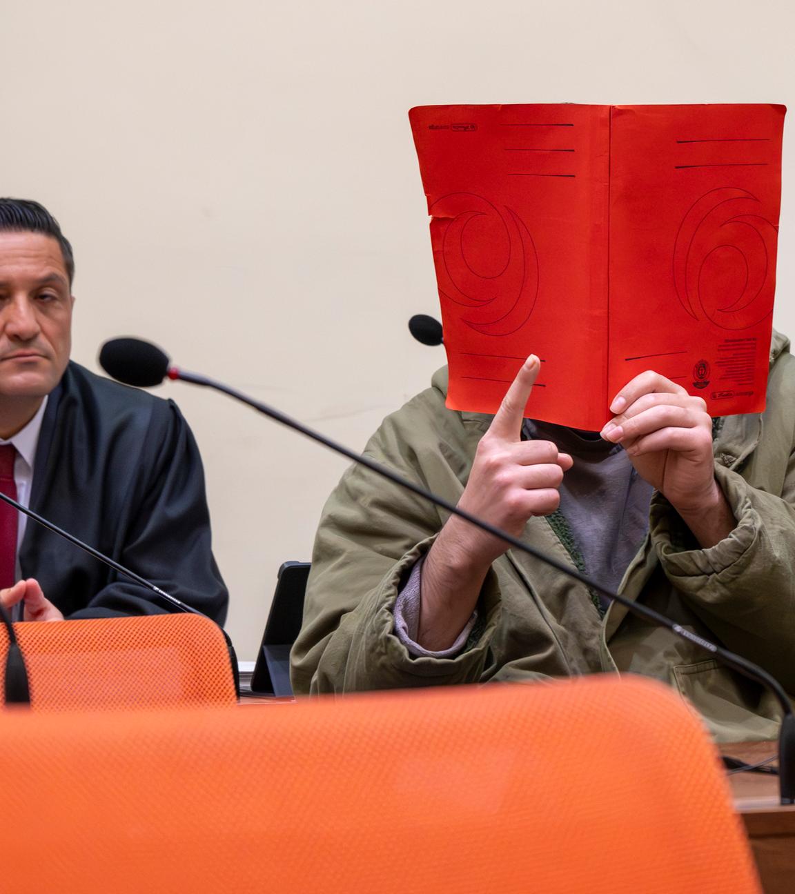  Der Angeklagte Farhad N. sitzt zu Prozessbeginn im Prozesssaal mit erhobenen rechten Zeigefinger neben seinem Anwalt Ömer Sahinci.