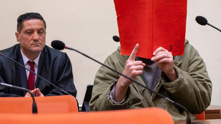  Der Angeklagte Farhad N. sitzt zu Prozessbeginn im Prozesssaal mit erhobenen rechten Zeigefinger neben seinem Anwalt Ömer Sahinci.