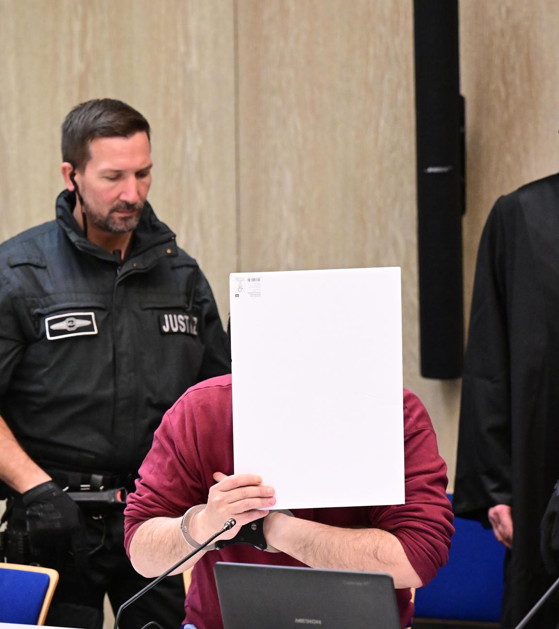  Der Angeklagte in einem Mordprozess nach einer Todesfahrt von Mannheim sitzt mit einem Aktendeckel vor seinem Gesicht im Gerichtssaal, rechts neben ihm steht sein Verteidiger Uwe Kosmala.