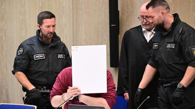  Der Angeklagte in einem Mordprozess nach einer Todesfahrt von Mannheim sitzt mit einem Aktendeckel vor seinem Gesicht im Gerichtssaal, rechts neben ihm steht sein Verteidiger Uwe Kosmala.