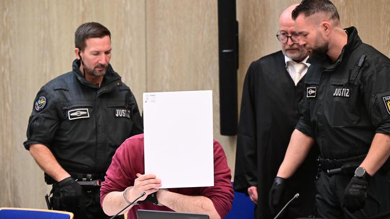  Der Angeklagte in einem Mordprozess nach einer Todesfahrt von Mannheim sitzt mit einem Aktendeckel vor seinem Gesicht im Gerichtssaal, rechts neben ihm steht sein Verteidiger Uwe Kosmala.