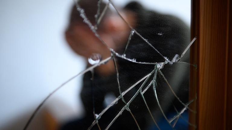 Ein Mann sitzt neben einem gesprungenen Spiegel auf einem Bett und hält seinen Kopf in den Händen (gestellte Szene). Übermüdet, überlastet, erdrückt von angestautem Stress und gepeinigt durch unbewältigte Tiefs