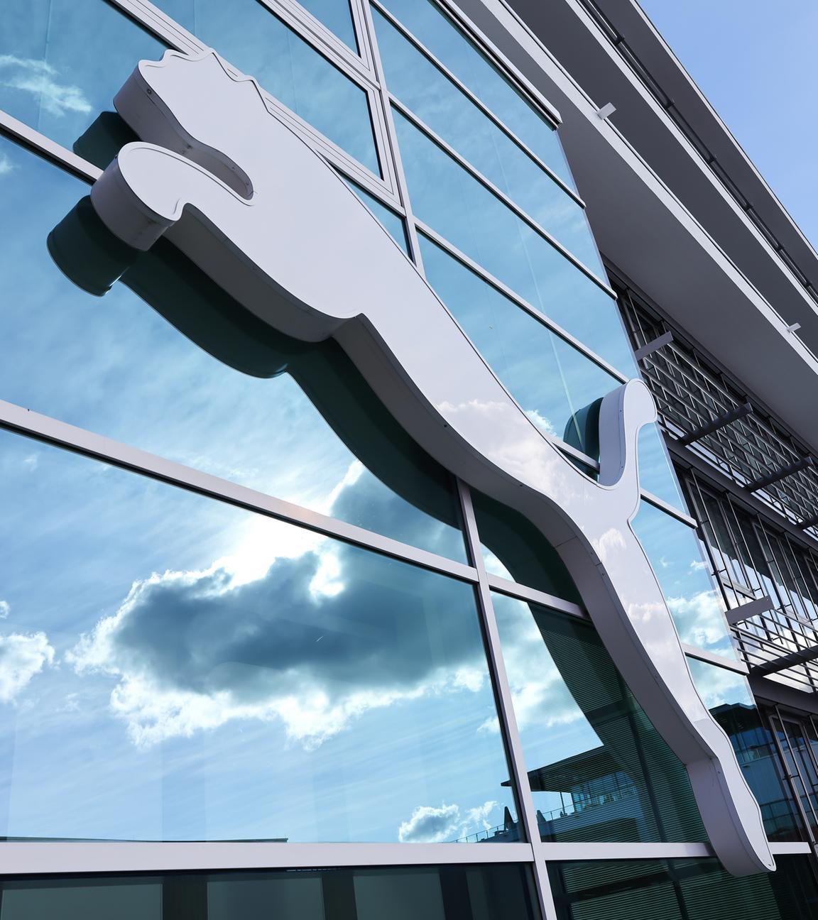 Bayern, Herzogenaurach: Wolken spiegeln sich im Fenster hinter einem Puma-Logo am Firmensitz des Sportartikelherstellers