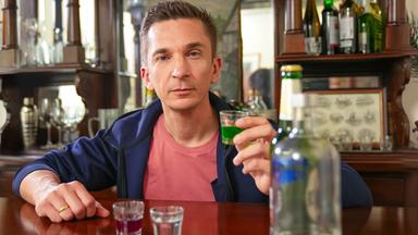 Eric sitzt an einer Bar und hält ein Glas mit Alkohol gefüllt in der Hand. Vor ihm stehen weitere Gläser und Flaschen.