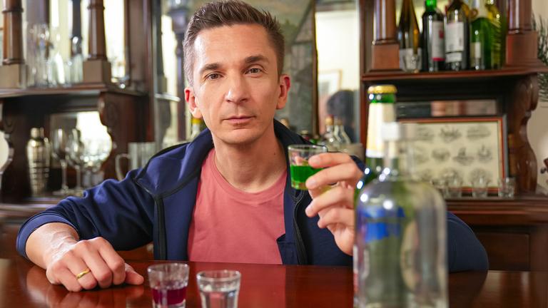 Eric sitzt an einer Bar und hält ein Glas mit Alkohol gefüllt in der Hand. Vor ihm stehen weitere Gläser und Flaschen.