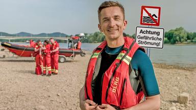 PUR+ Moderator Eric Mayer steht an einem sandigen Ufer des Flusses Rhein in einer roten Schwimmweste. Rechts neben ihm steht ein Schild mit dem Hinweis, dass das Baden hier wegen der Strömung verboten ist. Links neben ihm im Hintergrund sieht man mehrere Rettungsschwimmer an einem kleinen Rettungsboot.