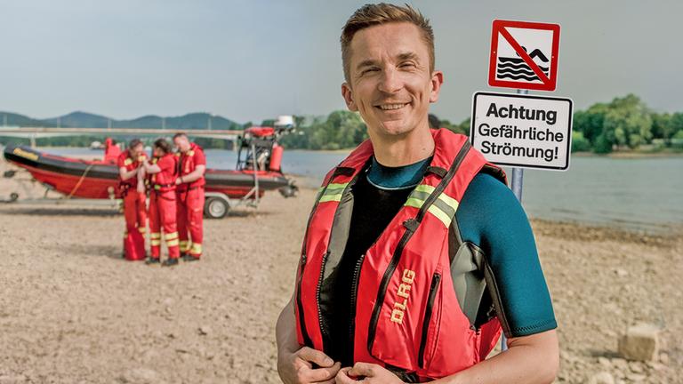 PUR+ Moderator Eric Mayer steht an einem sandigen Ufer des Flusses Rhein in einer roten Schwimmweste. Rechts neben ihm steht ein Schild, dass darauf hinweist, dass das Baden hier wegen der Strömung verboten ist. Links neben ihm im Hintergrund sieht man mehrere Rettungsschwimmer an einem kleinen Rettungsboot.