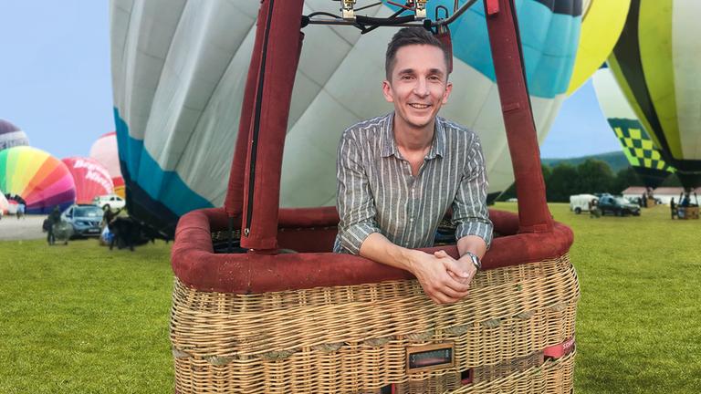 PUR+ Moderator Eric Mayer steht im Korb eines Heißluftballons, der noch auf der Erde steht. Wir sehen Eric relativ nah. Im Hintergrund liegen auf einer Wiese noch andere Ballone, die gerade aufgeblasen werden.