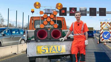 PUR+ Moderator Eric Mayer steht in orangefarbener Arbeitskleidung am Heck eines Baustellenfahrzeugs. Dieses hat auf der Ladefläche ein großes Warnkreuz mit Signallampen und ein Schild mit dem Schriftzug "Stau".
