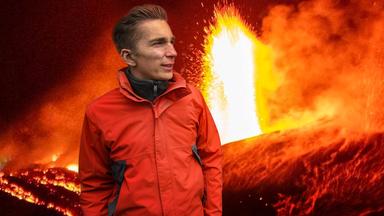 Eric steht vor einem ausbrechenden Vulkan, es spritzt Feuer in einer Fontäne nach oben (Collage).
