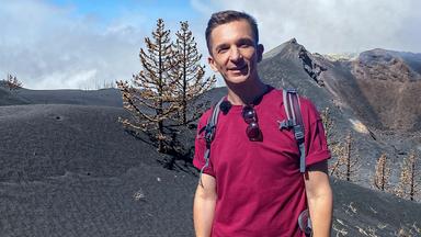 Eric schaut frontal in die Kamera. Hinter ihm sind schwarze Hügel und Berge und vertrocknet aussehende Bäume.