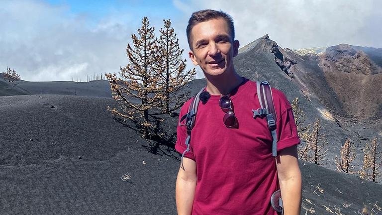 Eric schaut frontal in die Kamera. Hinter ihm sind schwarze Hügel und Berge und vertrocknet aussehende Bäume.