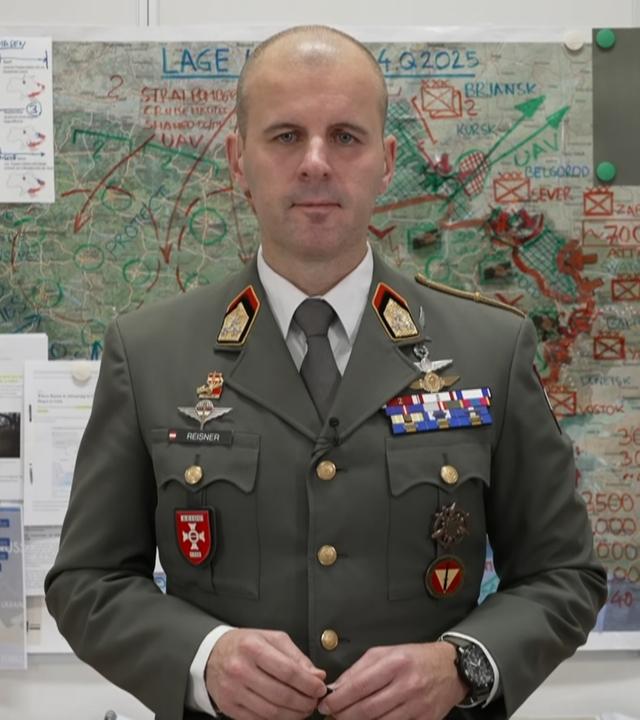 Oberst Markus Reisner stehend vor einer Karte an der Wand im Hintergrund.