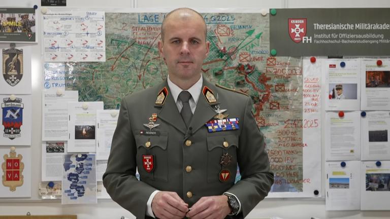 Oberst Markus Reisner stehend vor einer Karte an der Wand im Hintergrund.