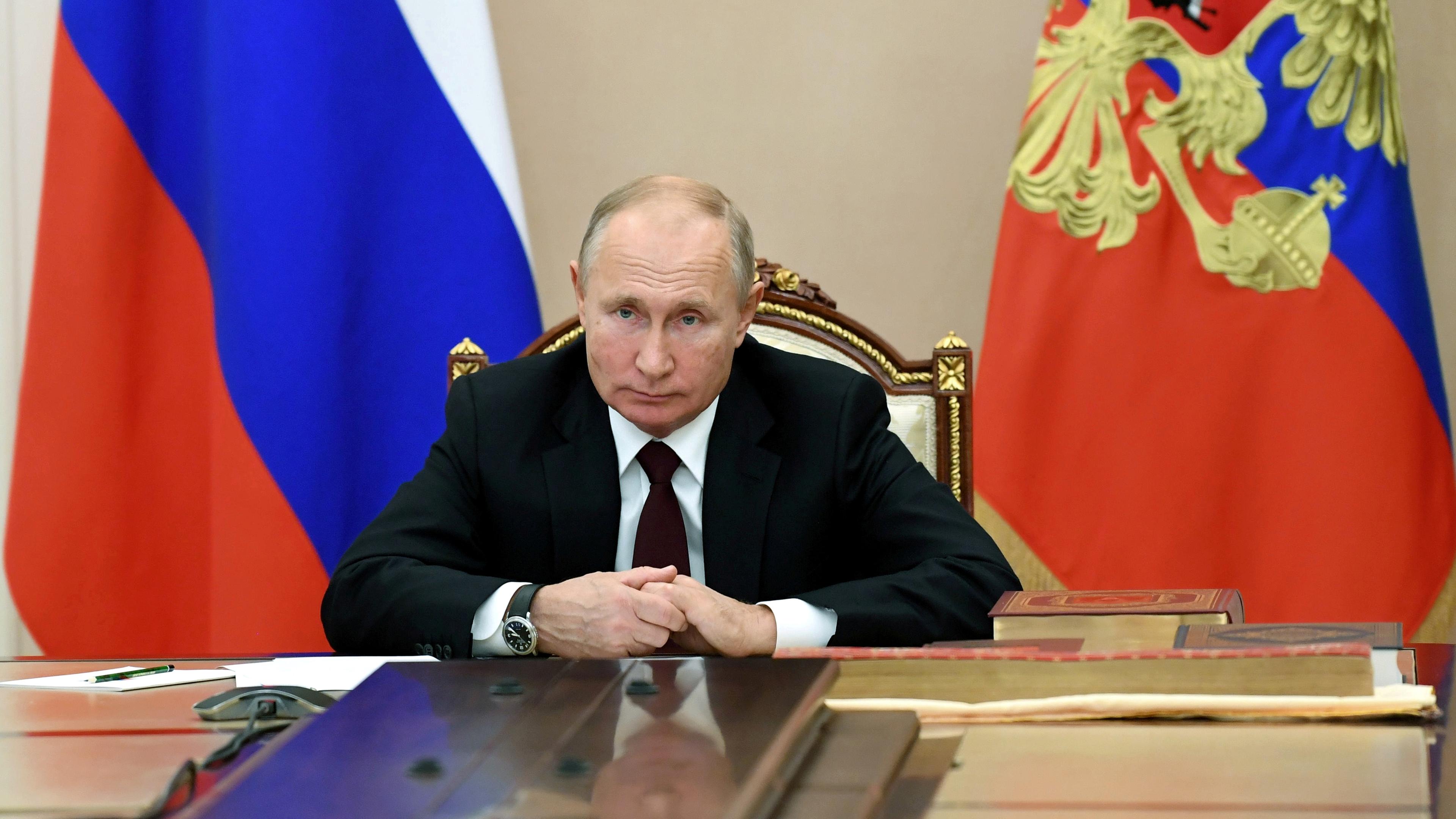 Wladimir Putin sitz an einem großen Tisch im Kreml, im Hintergrund russische Flaggen.