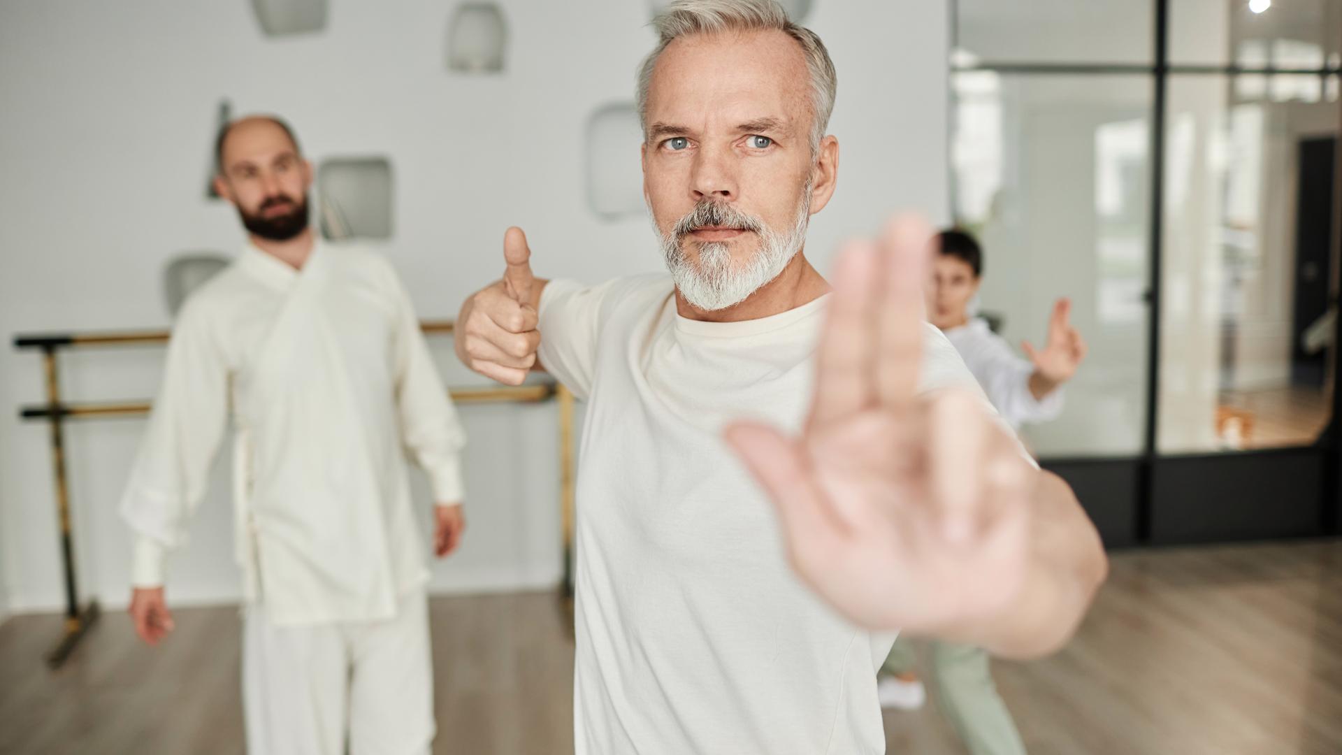Mann in einer Qigong-Pose, die von einem Trainer im Hintergrund begutachtet wird.
