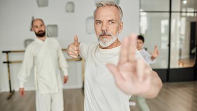 Mann in einer Qigong-Pose, die von einem Trainer im Hintergrund begutachtet wird.