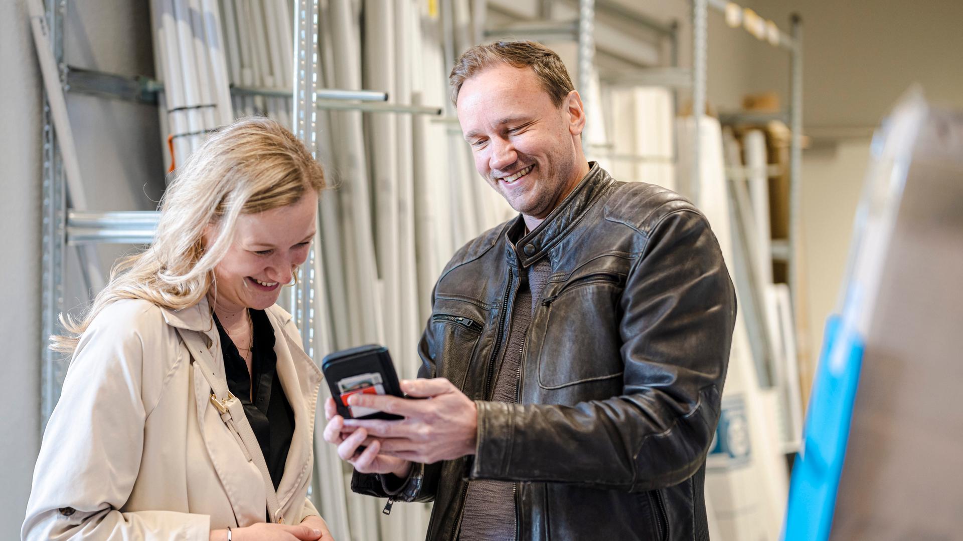 Es sind eine Frau und Mann zu sehen, diese stehen in einem handwerklichen Lager und sprechen miteinander.