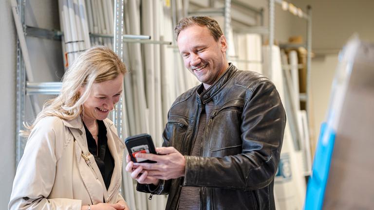 Es sind eine Frau und Mann zu sehen, diese stehen in einem handwerklichen Lager und sprechen miteinander.