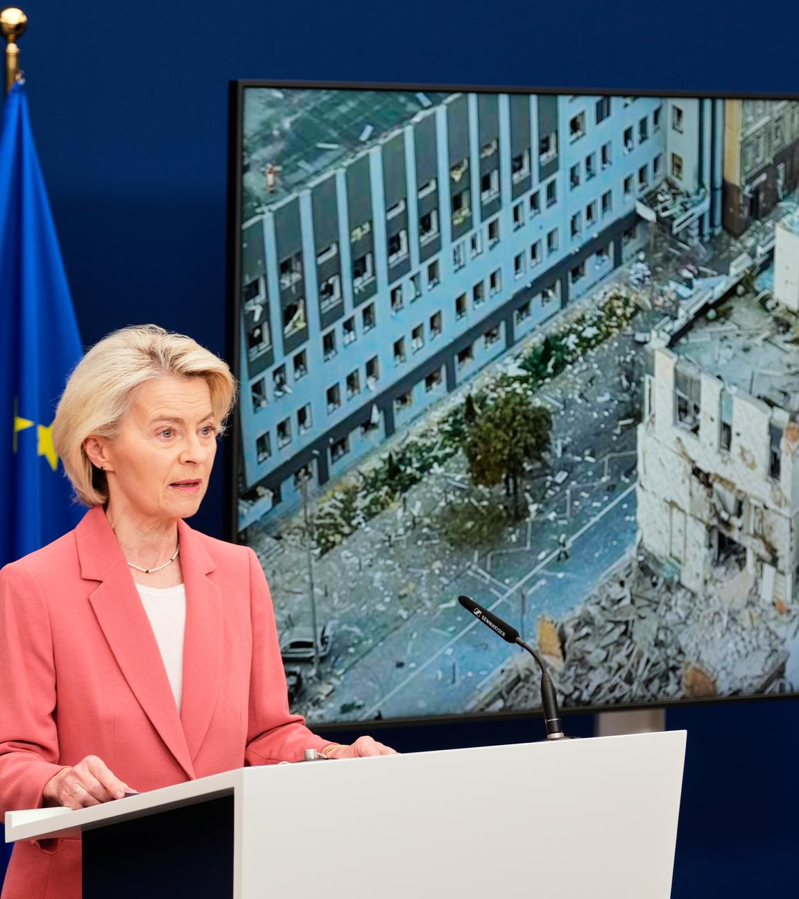 Ursula von der Leyen steht an einem Rednerpult. Hinter ihr ist ein Foto des beschädigten Geäudes der EU-Vertretung. Außerdem steht hinter ihr eine eurpäische Flagge.
