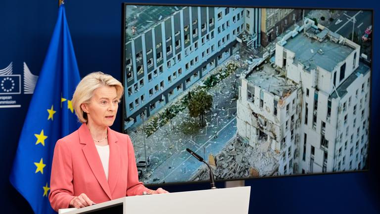 Ursula von der Leyen steht an einem Rednerpult. Hinter ihr ist ein Foto des beschädigten Geäudes der EU-Vertretung. Außerdem steht hinter ihr eine eurpäische Flagge.
