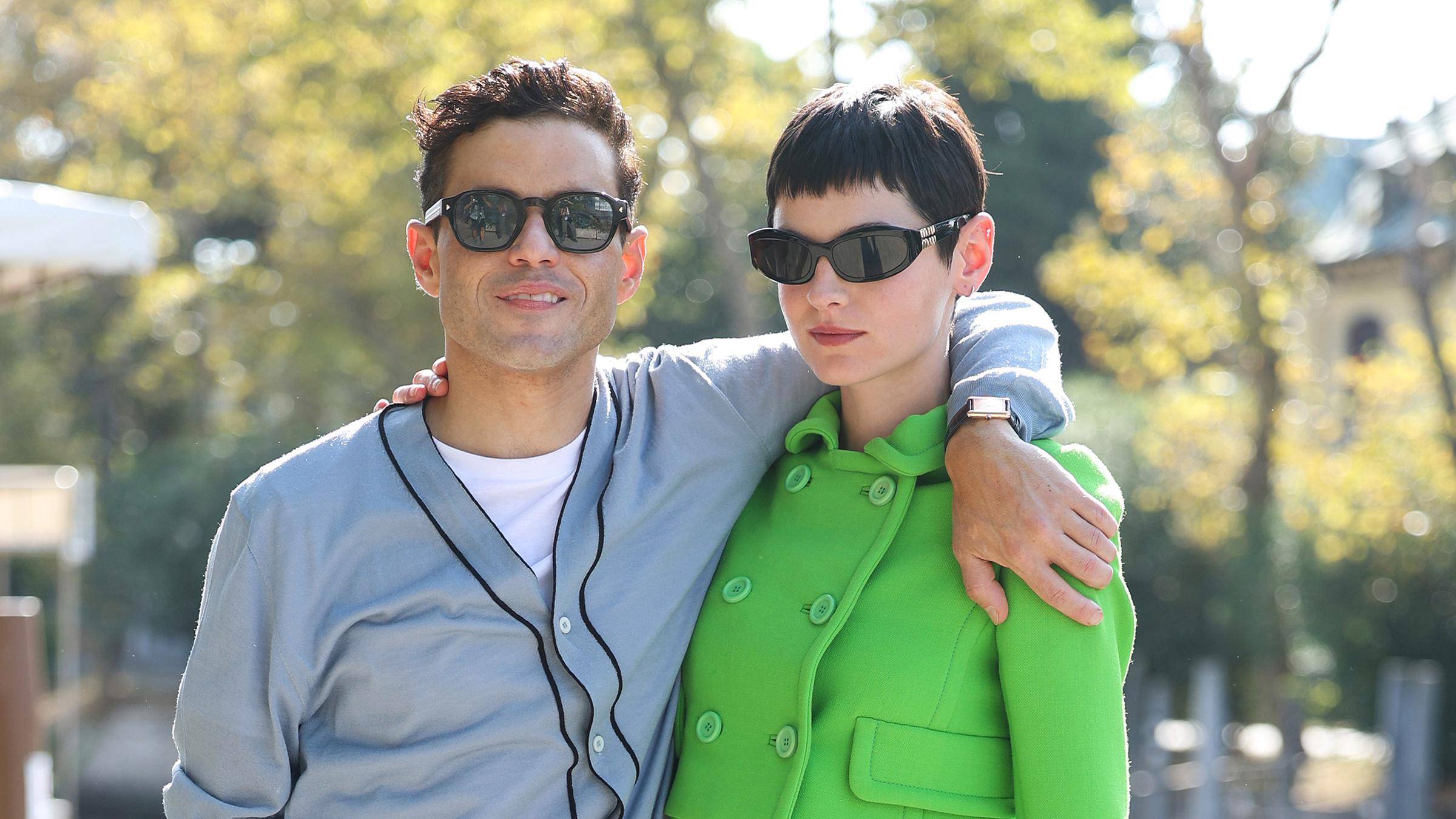 Rami Malek in einem grauen und Emma Corrin in einem grünen Oberteil posieren für ein gemeinsames Foto. Beide tragen Sonnenbrillen und legen einen Arm um die Schulter des anderen.