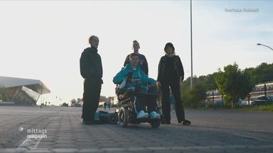 Drei Männer stehen lässig auf der Straße, einer sitzt im Rollstuhl davor