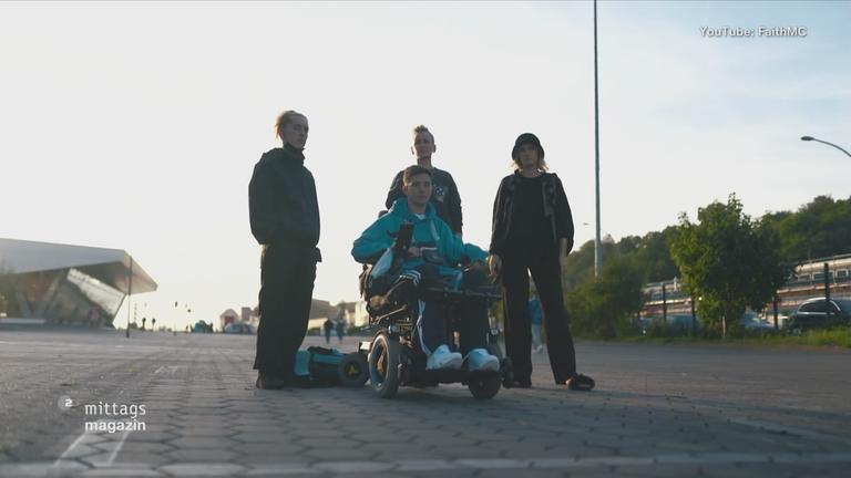 Drei Männer stehen lässig auf der Straße, einer sitzt im Rollstuhl davor