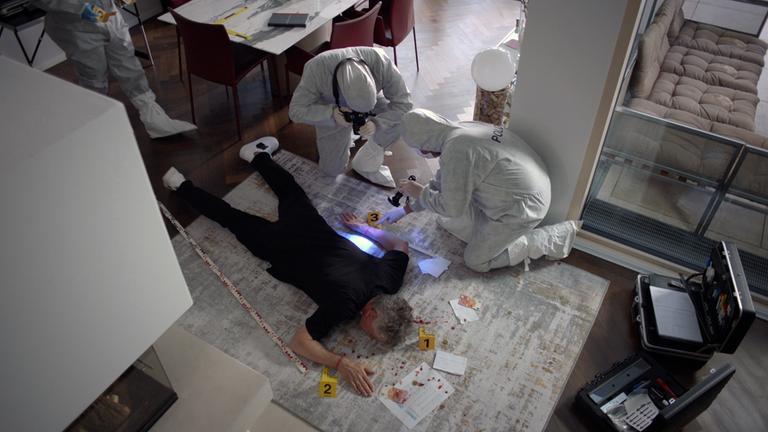 Nachgestellte Szene: Ein Mann liegt tot auf einem Teppich. Um ihn herum knien mehrere Personen in Schutzanzügen der Polizei. Sie untersuchen die Leiche.