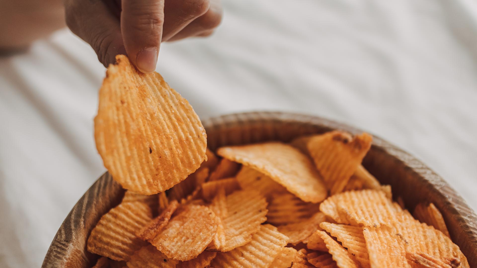 Eine Hand greift aus einer Schale mit Kartoffelchips einen Cjip heraus. 
