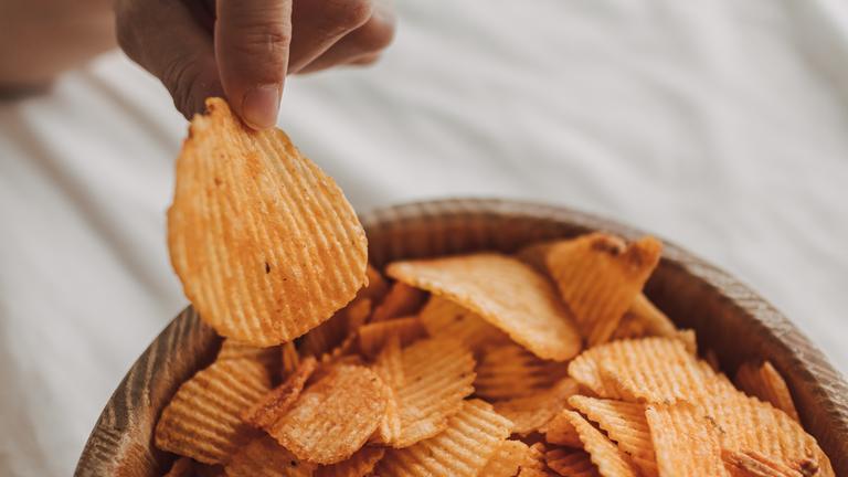 Eine Hand greift aus einer Schale mit Kartoffelchips einen Cjip heraus. 
