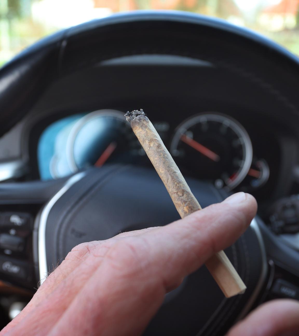Ein Mann sitzt mit einem Joint zwischen den Fingern am Steuer eines Autos.