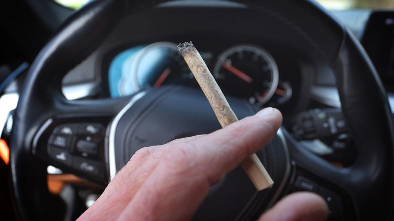 Ein Mann sitzt mit einem Joint zwischen den Fingern am Steuer eines Autos.