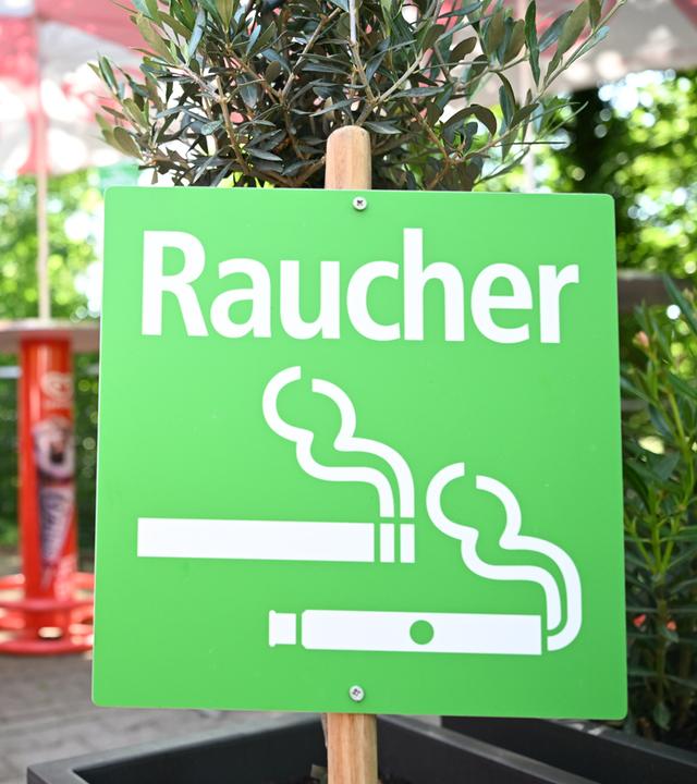 Eine Frau steht rauchend in einem ausgewiesenen Raucherbereich in einem Freibad neben einem grünen Hinweisschild mit der Aufschrift "Raucher" am 27.05.2026 in Stuttgart.