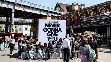 Menschen sitzen und stehen um ein Plakat der re:publica mit dem Mottospruch. Im Hintergrund ein Gebäude bei sonnigem Wetter.