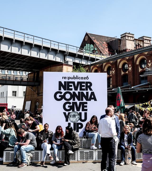Menschen sitzen und stehen um ein Plakat der re:publica mit dem Mottospruch. Im Hintergrund ein Gebäude bei sonnigem Wetter.