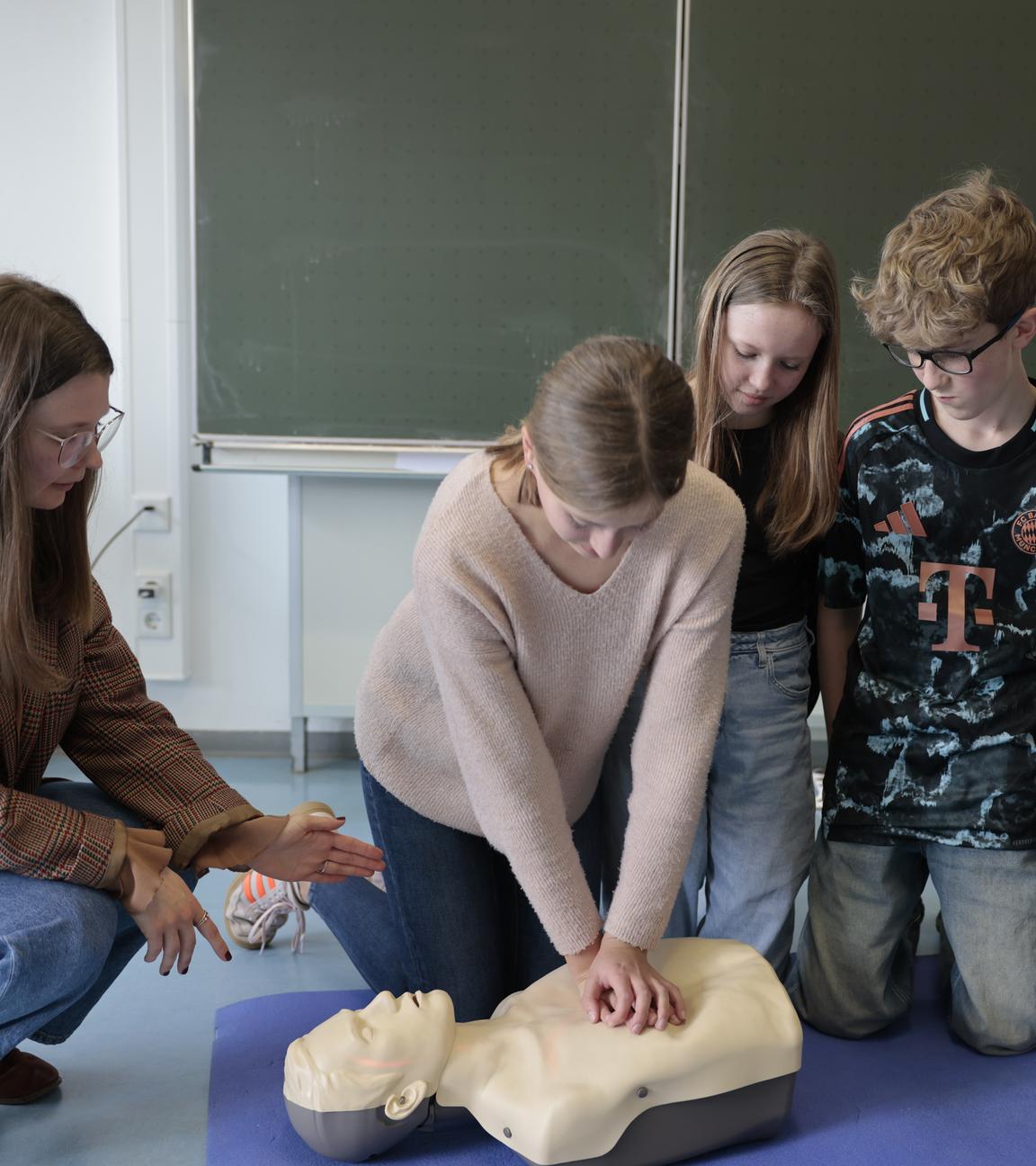 Eine Frau kniet mit zwei Schülern und zwei Schülerin vor einer Trainingspuppe. Eine Schülerin hat ihre Hände auf dem Brustkorb der Puppe gelegt.
