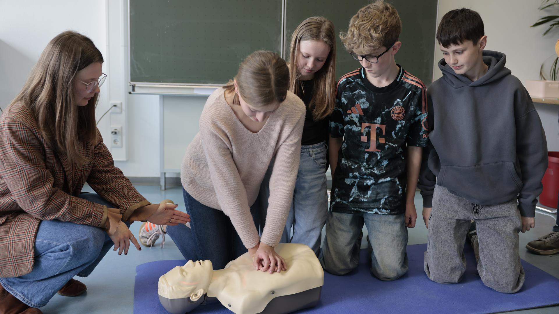 Eine Frau kniet mit zwei Schülern und zwei Schülerin vor einer Trainingspuppe. Eine Schülerin hat ihre Hände auf dem Brustkorb der Puppe gelegt.