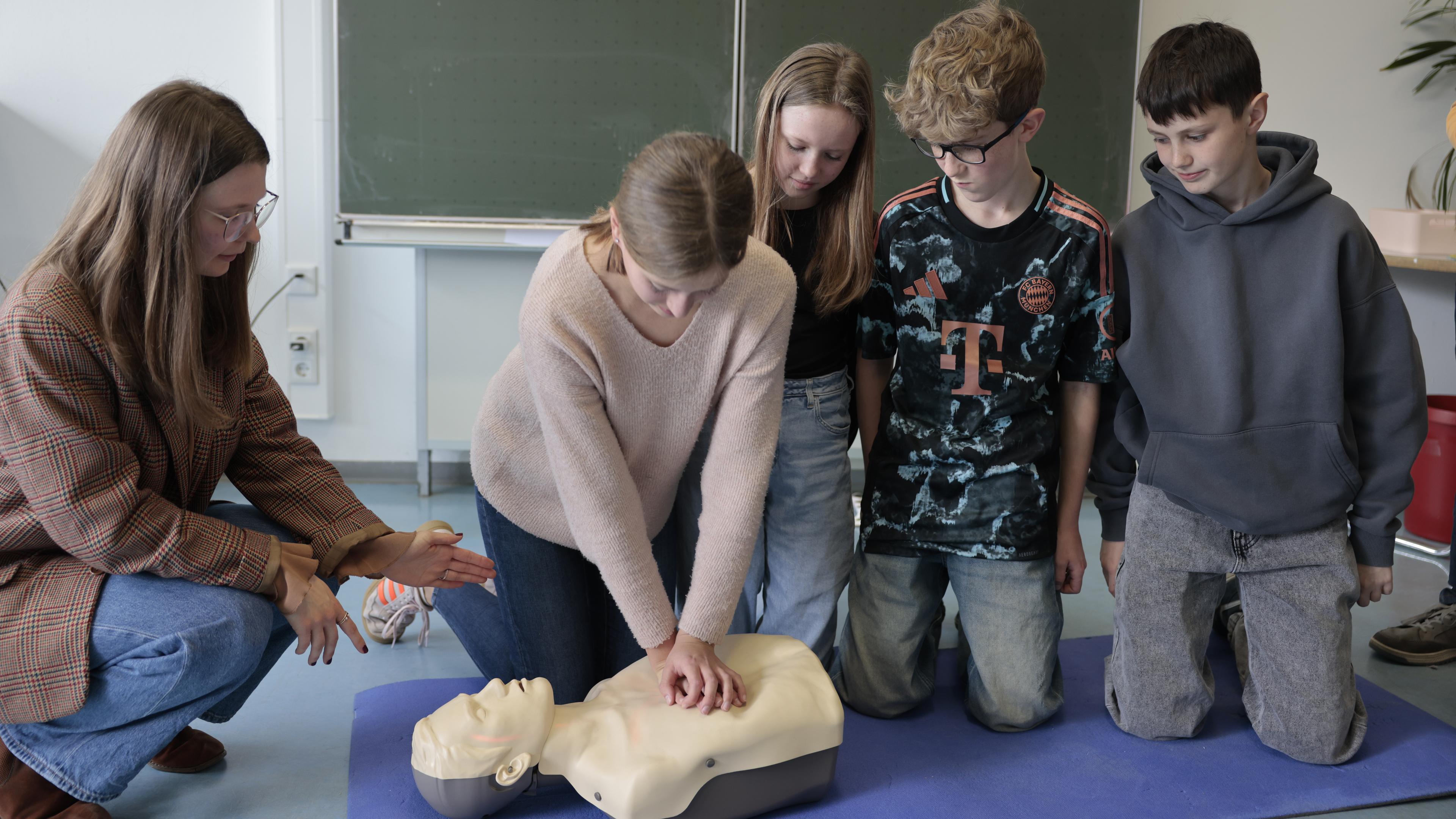 Eine Frau kniet mit zwei Schülern und zwei Schülerin vor einer Trainingspuppe. Eine Schülerin hat ihre Hände auf dem Brustkorb der Puppe gelegt.