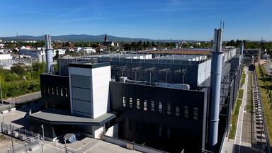 Luftaufnahme eines großen umzäunten Gebäudes mit schwarzer Fassade. Dahinter die Dächer von Hattersheim.
