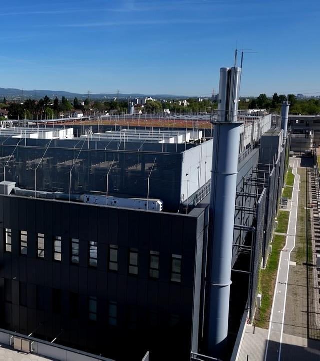 Luftaufnahme eines großen umzäunten Gebäudes mit schwarzer Fassade. Dahinter die Dächer von Hattersheim.