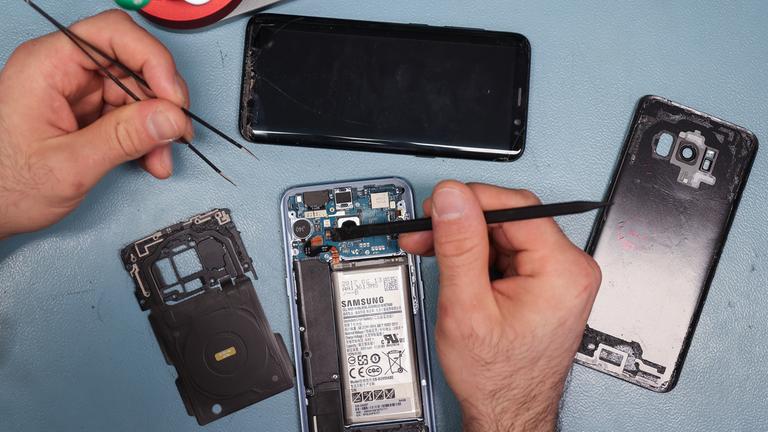 Zwei Hände halten Werkzeuge, auf der Oberfläche dahinter liegen zum Teil auseinander gebaute Smartphones.