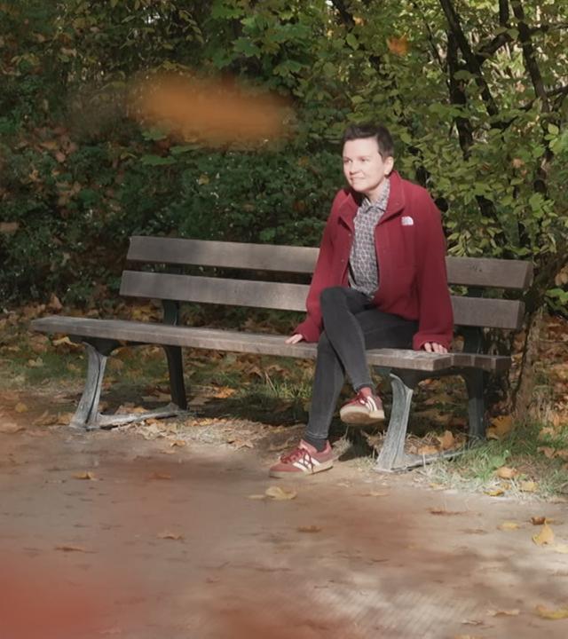 Zu sehen ist Eva-Maria Wild auf einer Bank in einem Park. 