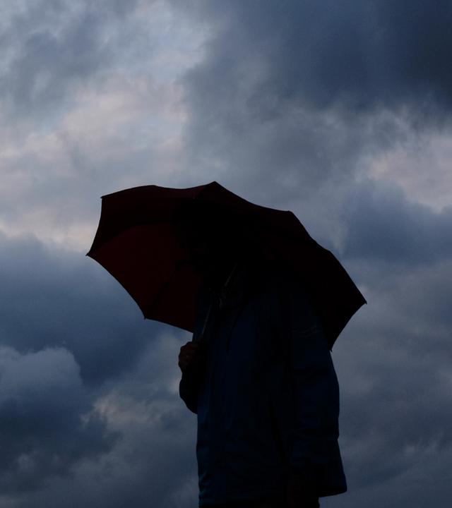Ein Mann geht mit einem Regenschirm vor aufziehenden dunklen Wolken spazieren. (Archiv)