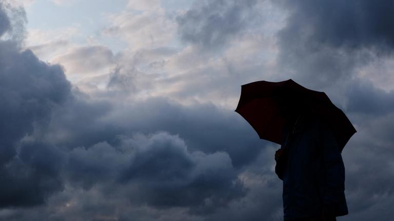 Ein Mann geht mit einem Regenschirm vor aufziehenden dunklen Wolken spazieren. (Archiv)