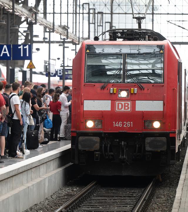 Fahrgäste warten dicht gedrängt an einem Bahnsteig. Ein Zug steht auf dem Gleis.