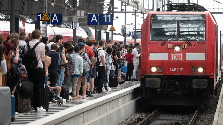 Fahrgäste warten dicht gedrängt an einem Bahnsteig. Ein Zug steht auf dem Gleis.