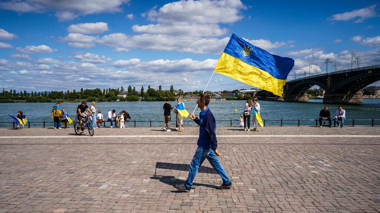 Archiv: Ein Mann geht auf einer Kundgebung zum 34. Unabhängigkeitstag der Ukraine mit der ukrainischen Nationalflagge am Rheinufer.